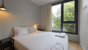 A hotel room with a large bed, a bedside table, a nightstand, a window with curtains, and a hanging light fixture. - FLH Boavista Flat II (Oporto)