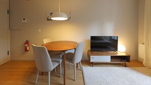 A modern living room with a wooden dining table, grey chairs, a flat-screen TV, a white pendant light, and a fire extinguisher on the wall. - FLH Boavista Flat II (Oporto)