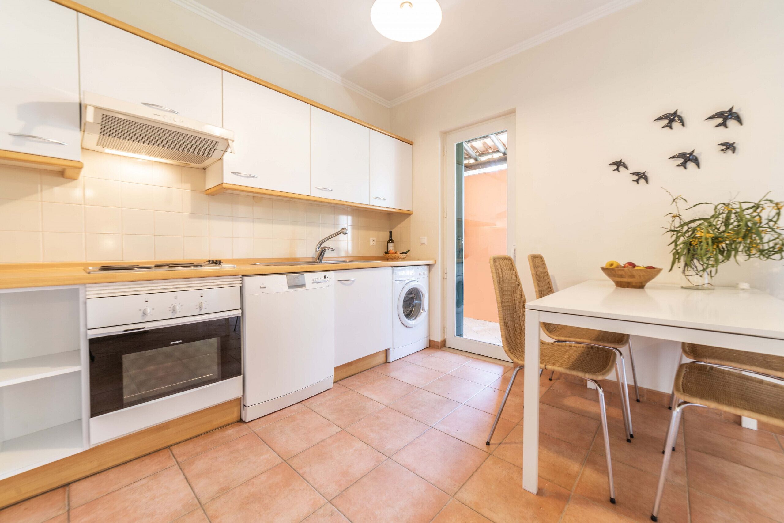 Une cuisine moderne avec des armoires blanches, un plan de travail en bois, un four, un lave-vaisselle et un lave-linge. On y trouve une table à manger avec des chaises et un vase rempli de fleurs.