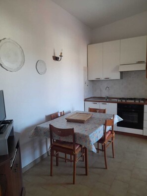 Una cocina compacta con armarios blancos, piso de baldosas, una mesa de comedor con sillas y una estufa pequeña.