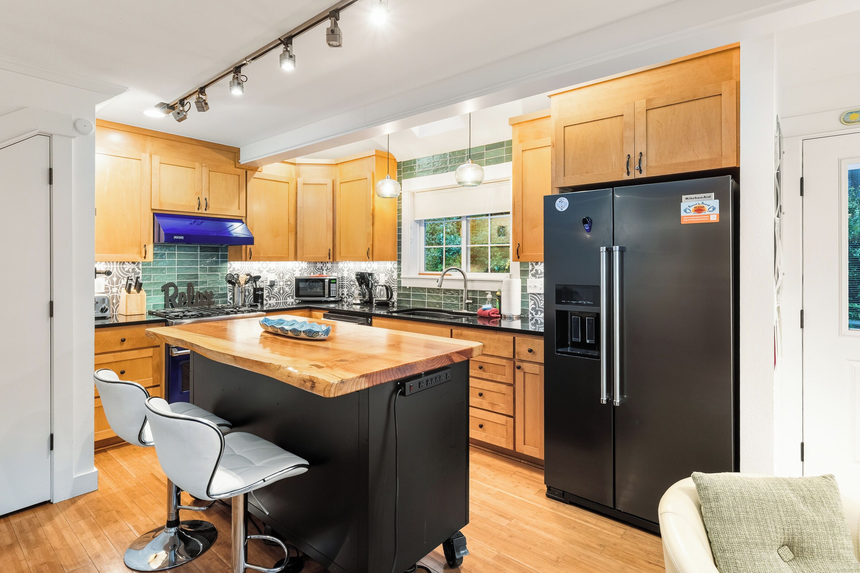 Una cocina moderna con armarios de madera, un refrigerador negro, una isla de madera con taburetes de bar y una ventana que proporciona luz natural.