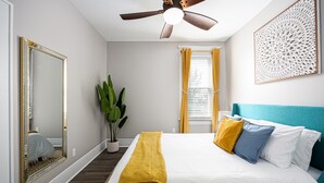 Un dormitorio con cama, ventilador de techo, espejo grande, planta en maceta y ventana con cortinas.