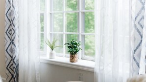 A window with a view of greenery, a white wall, and a patterned curtain.