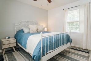 Ein ordentlich eingerichtetes Schlafzimmer mit weißem Metallbettgestell, einer blauen und weißen Bettdecke, einem Nachttisch mit Lampe und einem Fenster mit Vorhängen.