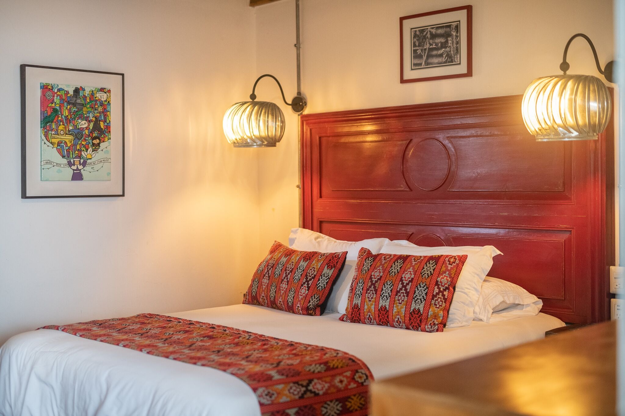 Habitación de hotel con una cama grande, dos almohadas estampadas, un cabecero rojo y dos lámparas de pared.