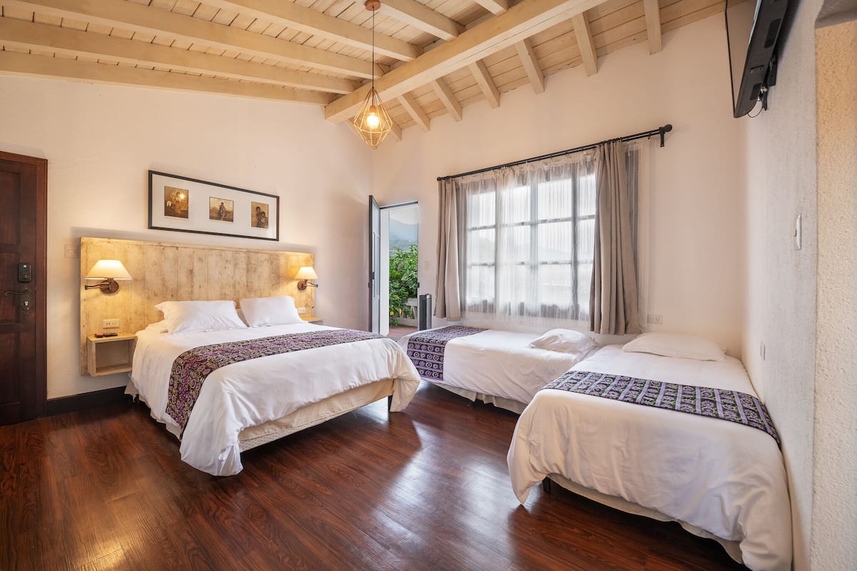 A hotel room with two beds, wooden flooring, a wooden ceiling, a large window with curtains, and a framed picture on the wall.