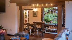 A dining area with a wooden table and chairs, a television mounted on the wall, and a view of a garden through an open doorway.