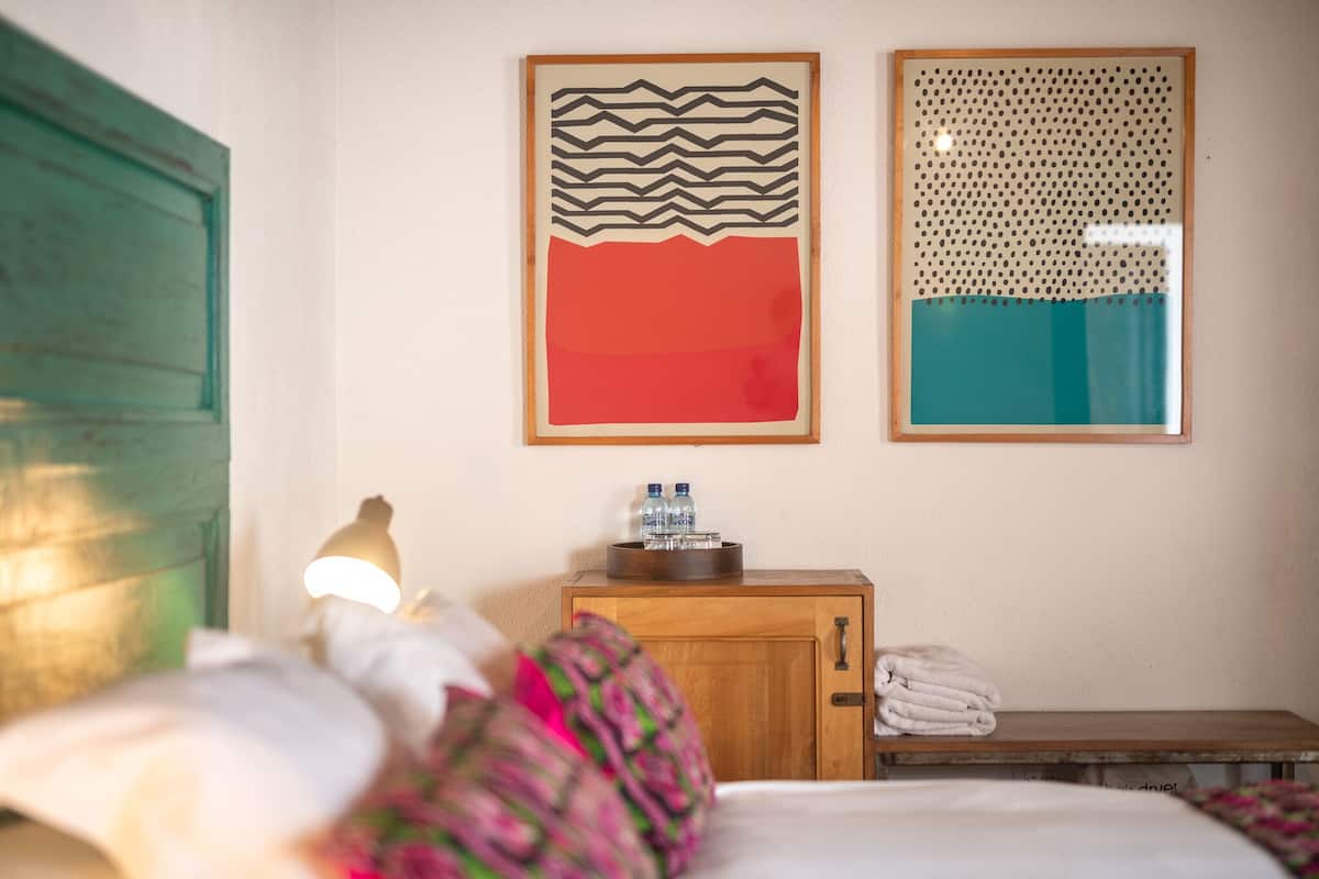 A hotel room with a bed, a wooden nightstand with a lamp, a wall with two framed artworks, and a window with a view of the outside.