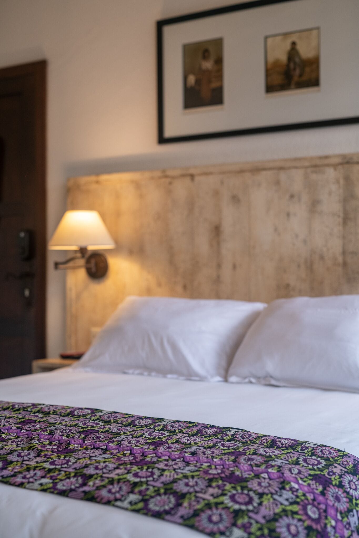 A bed with white linens and a floral-patterned blanket, a wooden headboard, a wall-mounted lamp, and two framed pictures above the bed.