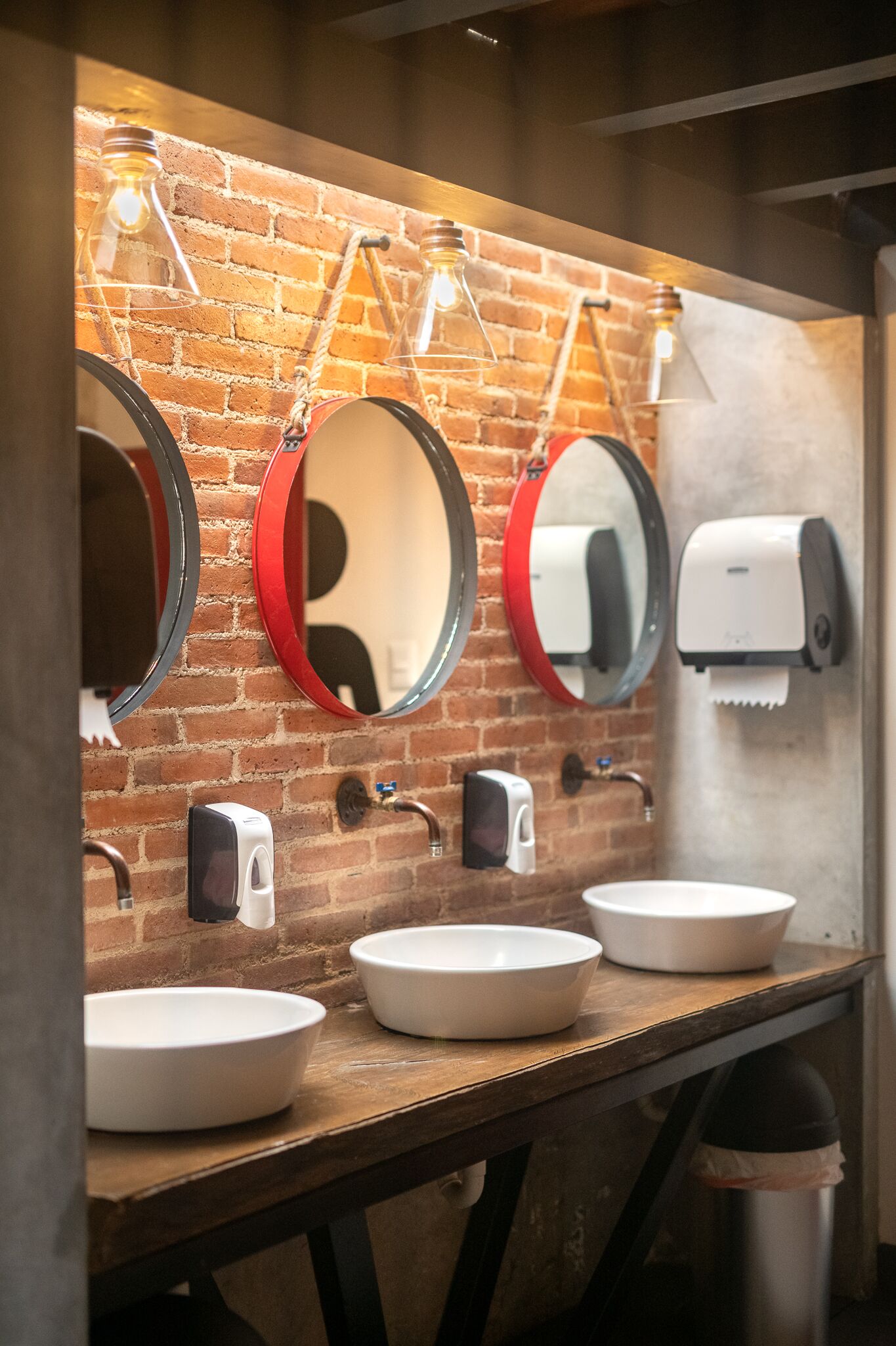 A restroom with three sinks, two round mirrors with red frames, and a brick wall.