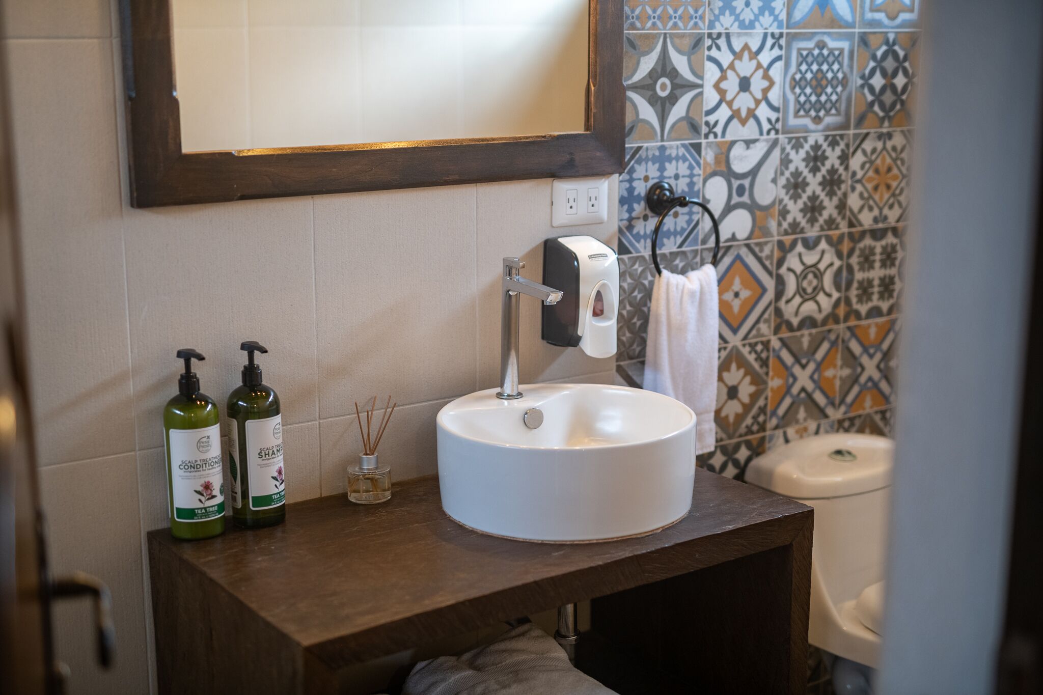 Un baño con un lavamanos blanco redondo, dos botellas verdes de champú, un pequeño frasco de vidrio con difusor de aroma y un espejo sobre el lavamanos.
