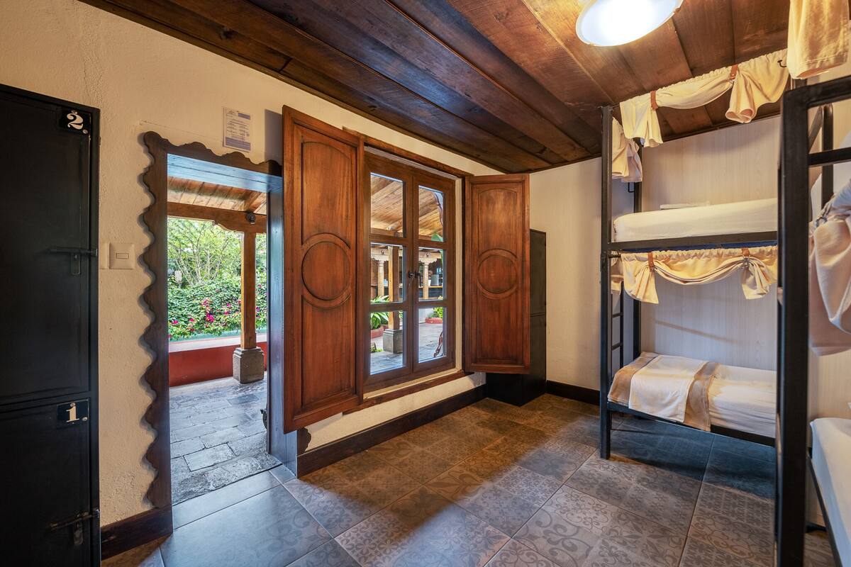 A room with a wooden ceiling, a window with curtains, a bunk bed, and a door with a number.