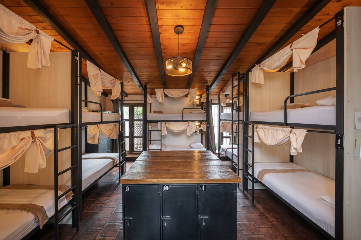 A dormitory-style room with bunk beds, a central desk, and a wooden ceiling.