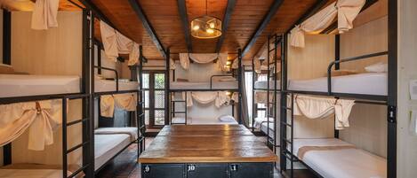 A dormitory-style room with bunk beds, a central desk, and a wooden ceiling.