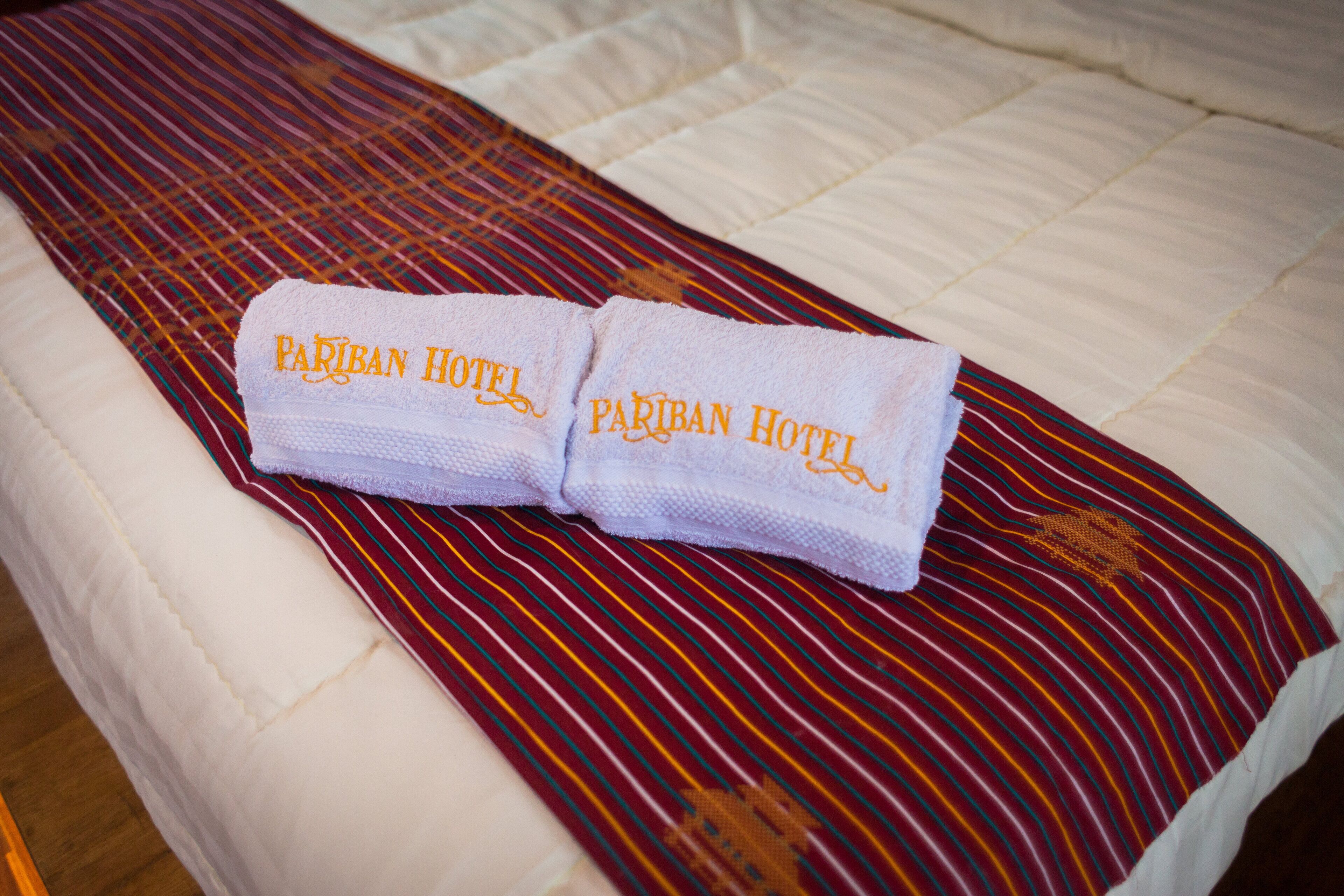 A bed with a white comforter and a striped red and yellow bedspread. Two folded towels with "PARIBAN HOTEL" embroidered on them are placed on the bed.