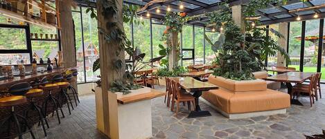 Un espacio interior amplio con techo de vidrio, suelo de piedra, muebles de madera y una zona de bar con taburetes.