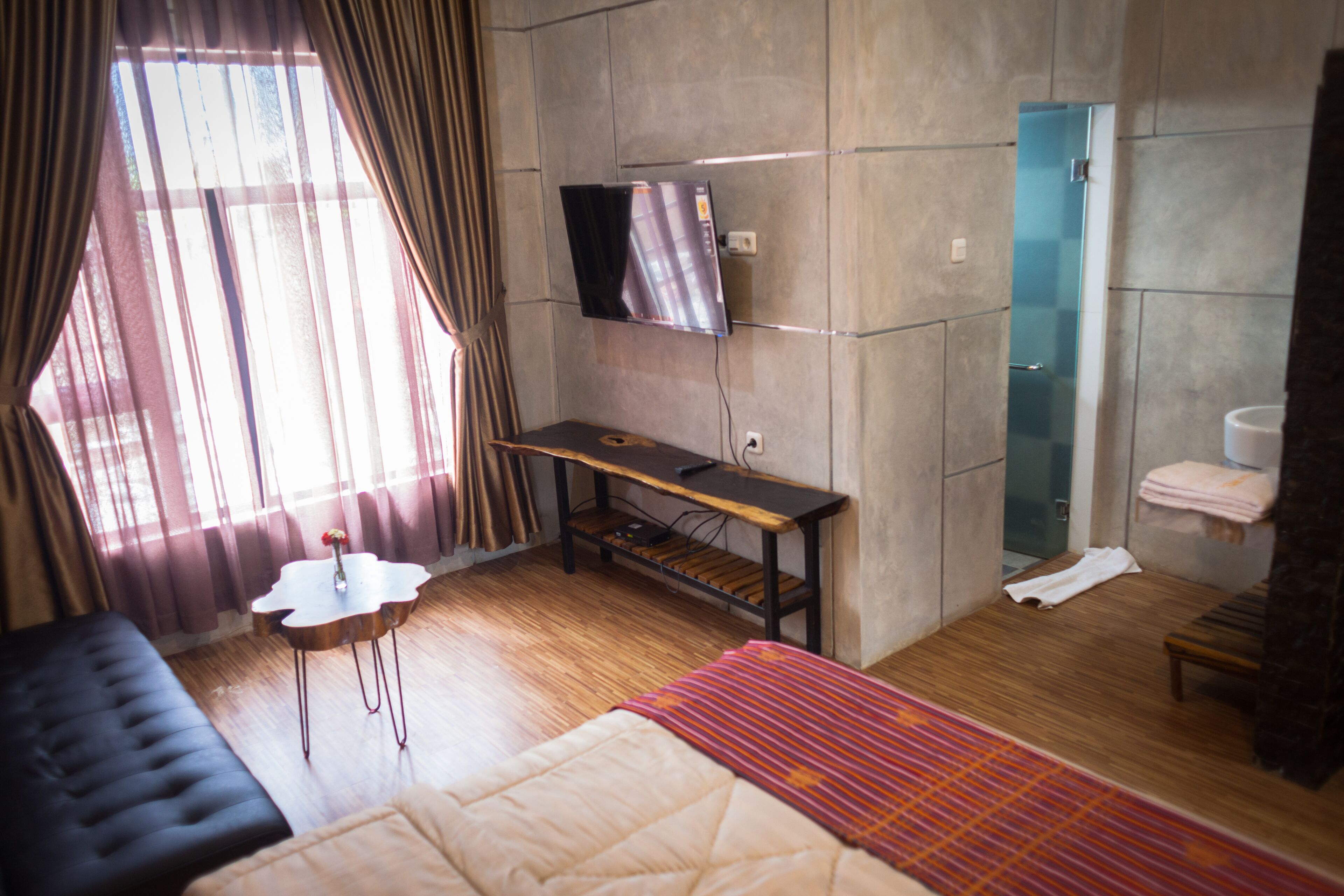 Quarto de hotel com cama, escrivaninha, TV fixada na parede, janela com cortinas e banheiro com porta-toalhas.