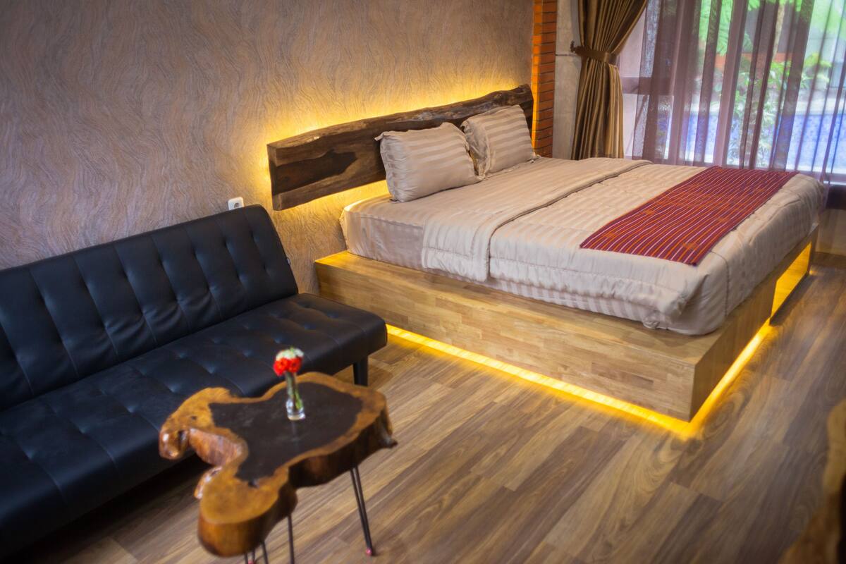 A bedroom with a bed, a black sofa, and a wooden coffee table with a guitar and a red flower vase.