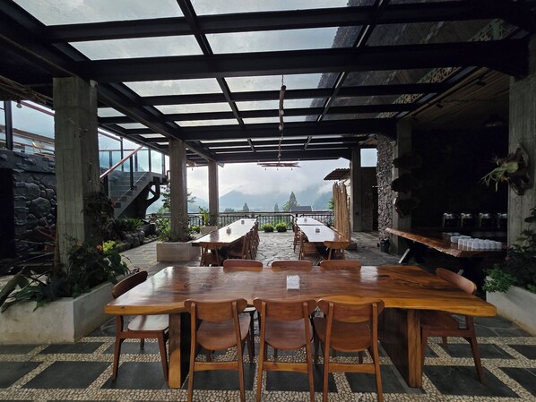 Un área de comedor al aire libre con una larga mesa de madera y sillas, rodeada de muros de piedra y plantas en macetas.