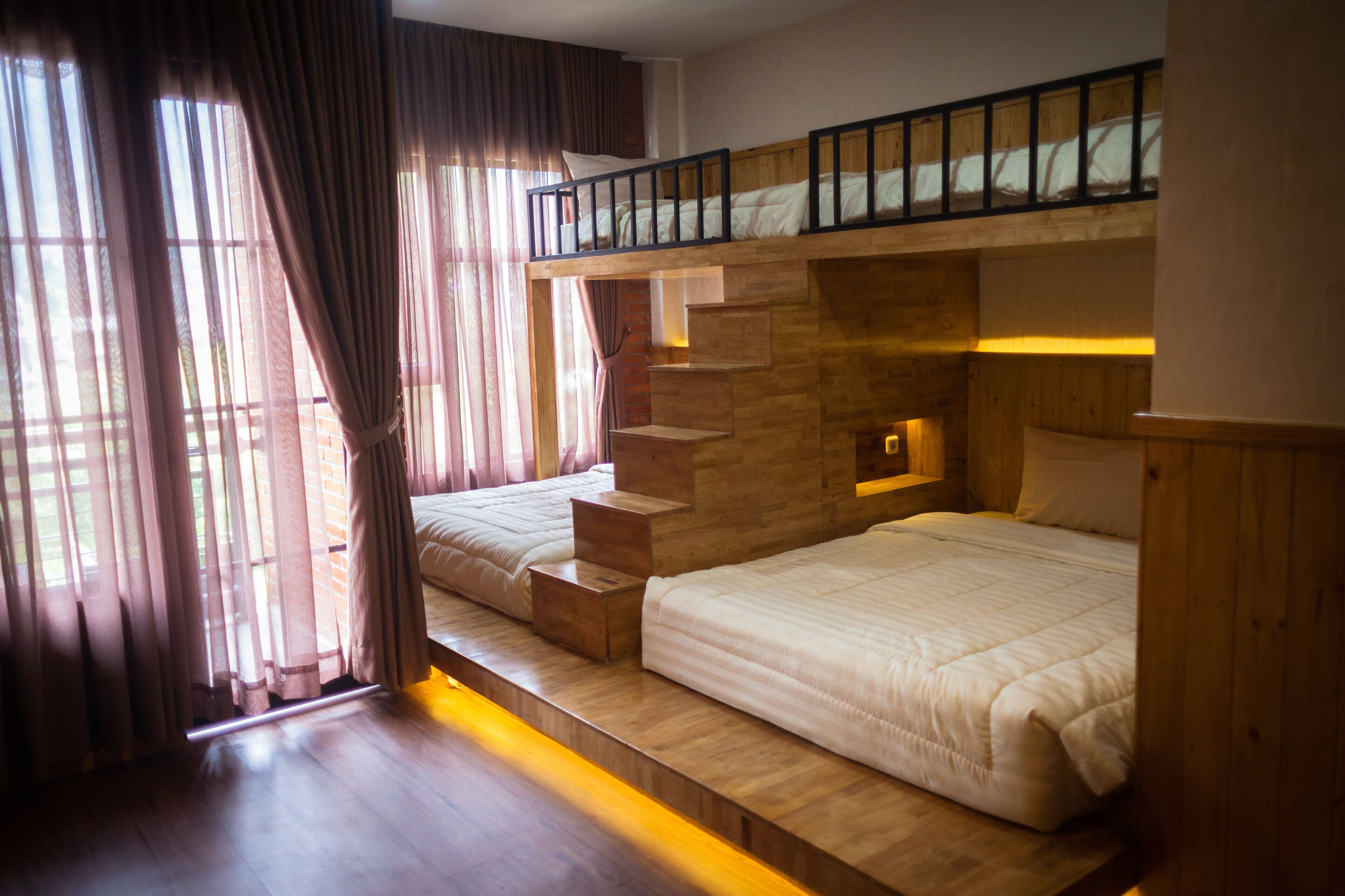 A room with two beds, wooden flooring, a staircase, and a window with curtains.