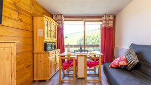 A cozy room with wooden walls, a dining table with chairs, a sofa, and a view of a green landscape. - L'Igloo - Studio pour 4 vue montagne (Villarembert)
