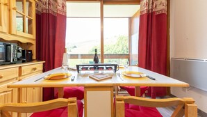 A dining area with a wooden table, red curtains, and a view of greenery outside. - L'Igloo - Studio pour 4 vue montagne (Villarembert)