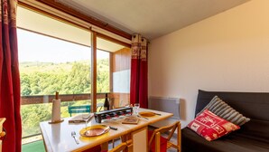 A room with a dining table set for two, a sofa with cushions, and a view of a forested landscape through a window. - L'Igloo - Studio pour 4 vue montagne (Villarembert)