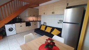 Uma cozinha compacta com máquina de lavar, uma mesa de jantar com um jogo americano vermelho, um sofá com almofadas amarelas e uma geladeira de aço inoxidável.