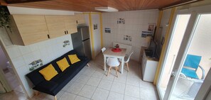 A compact kitchen with a dining area, a sofa with yellow cushions, a refrigerator, and a TV.