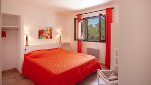 Ein Schlafzimmer mit einem großen Bett, einem Fenster mit roten Vorhängen, einem Nachttisch mit Lampe und einem Sessel.