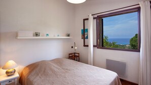Une chambre à coucher avec un lit, une table de chevet avec une lampe, une étagère avec des objets décoratifs, une fenêtre donnant sur la mer et un radiateur.