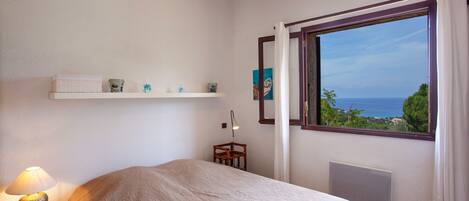 A bedroom with a bed, a nightstand with a lamp, a shelf with decorative items, a window with a view of the sea, and a radiator.