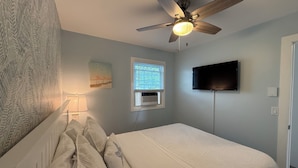 Une chambre à coucher comprenant un lit, un ventilateur de plafond, une fenêtre avec des stores, un téléviseur fixé au mur et un radiateur.