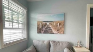 Une pièce avec un canapé gris, un tableau représentant un sentier de plage, une fenêtre avec des stores et une petite table de chevet agrémentée d’un objet décoratif.