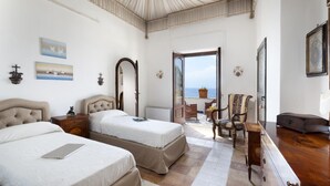 Quarto de hotel com duas camas, penteadeira de madeira, uma cadeira e vista para o mar através de uma porta de vidro.