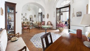 Ampla sala de estar com pé direito alto, mesa de jantar de madeira e diversos móveis antigos.