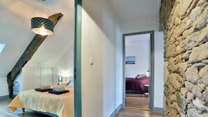 A bedroom with a bed, a wooden ceiling beam, a stone wall, and a view into another room.
