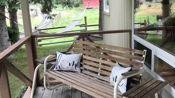 Een houten bank met witte kussens op een houten terras, met uitzicht op een gazon met bomen en een gebouw in rood in de verte.