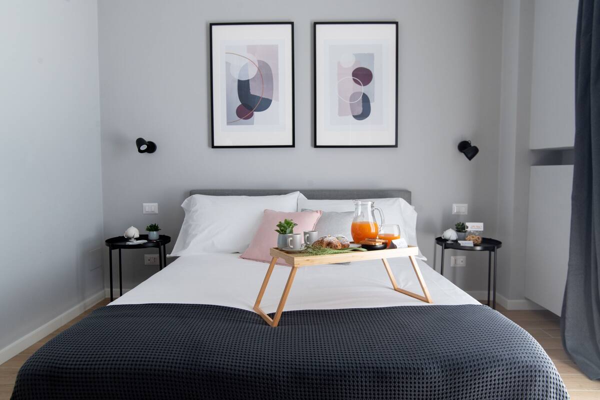 Een keurig opgemaakt bed met een dienblad ontbijtartikelen, een nachtkastje met een lamp, en twee ingelijste abstracte kunstwerken aan de muur.