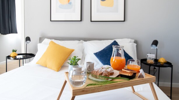 Een net opgemaakt bed met een dienblad vol ontbijtartikelen, zoals gebak, sap en water, in een moderne hotelkamer met kunst aan de muur.