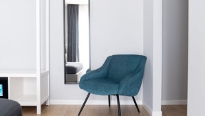 Een moderne kamer met een blauwgroene fauteuil, een witte boekenkast en een grote spiegel die een bed en gordijnen weerspiegelt.