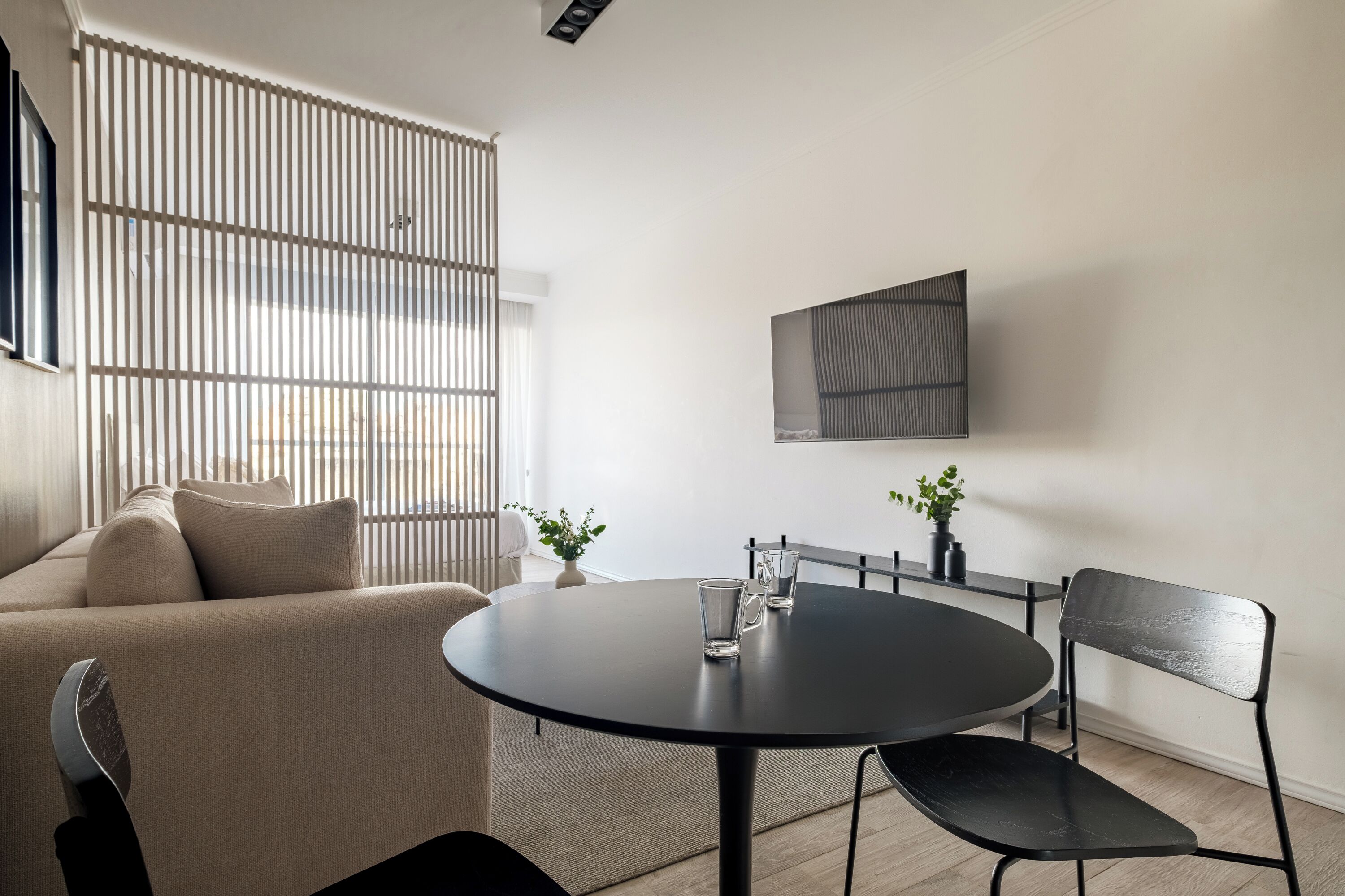 Ein modernes Wohnzimmer mit einem runden schwarzen Tisch, zwei schwarzen Sesseln, einem beigen Sofa und einem großen Fenster mit Jalousien.