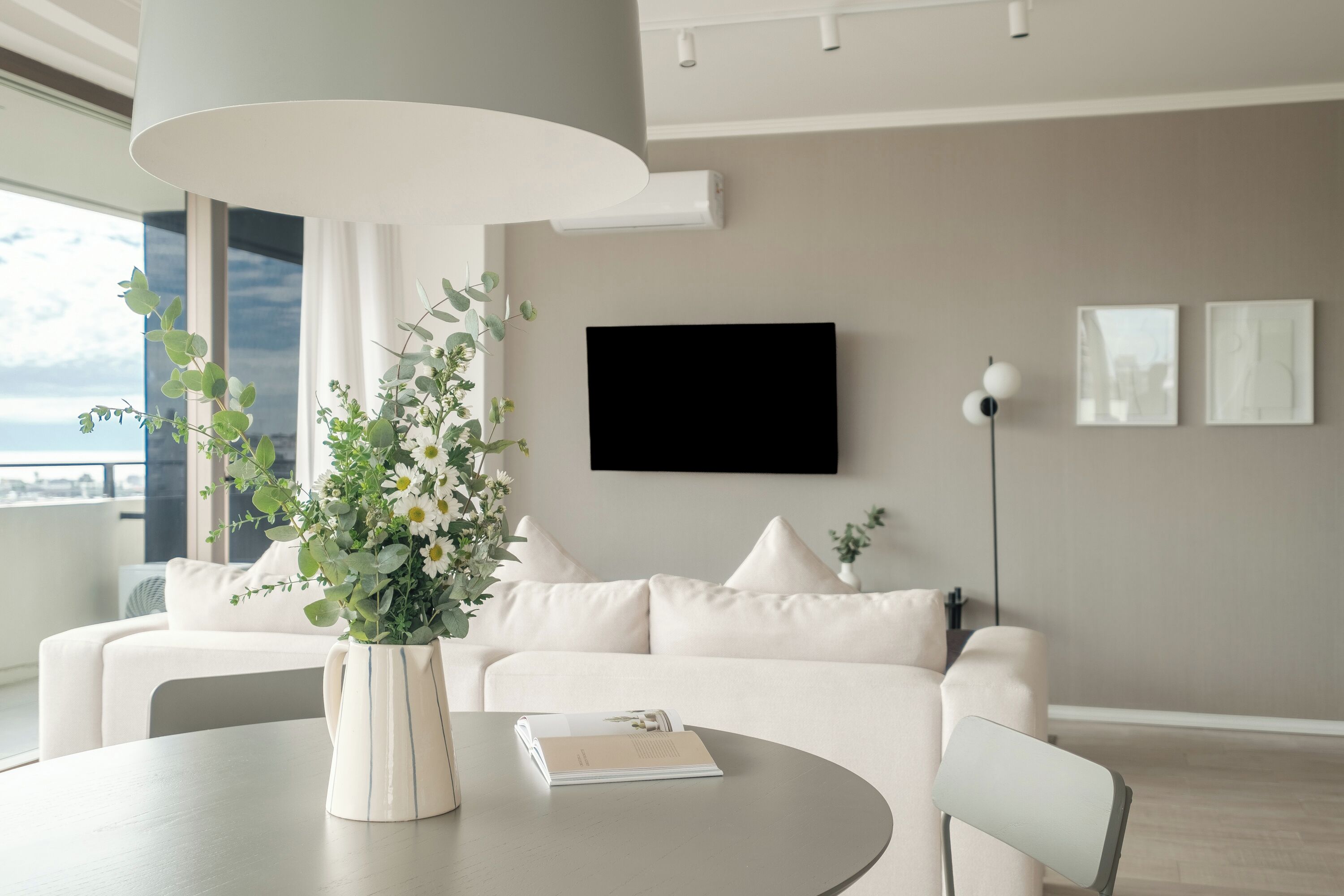 Un soggiorno moderno con un divano bianco, un tavolino con un vaso e un libro, un lampadario importante e una TV a schermo piatto sulla parete.