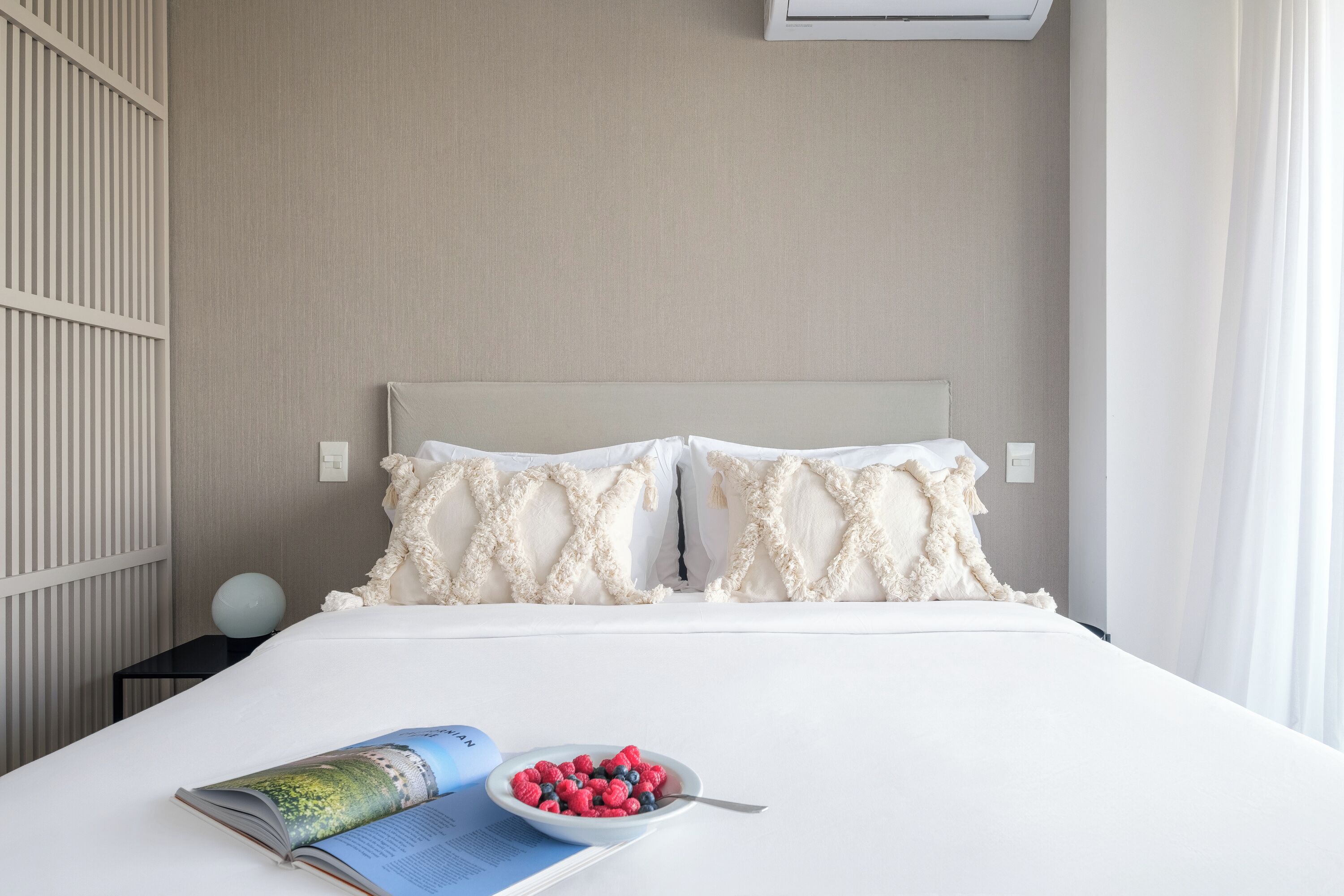 A neatly made bed with a magazine and a bowl of berries, a bedside lamp, and a wall-mounted air conditioner.