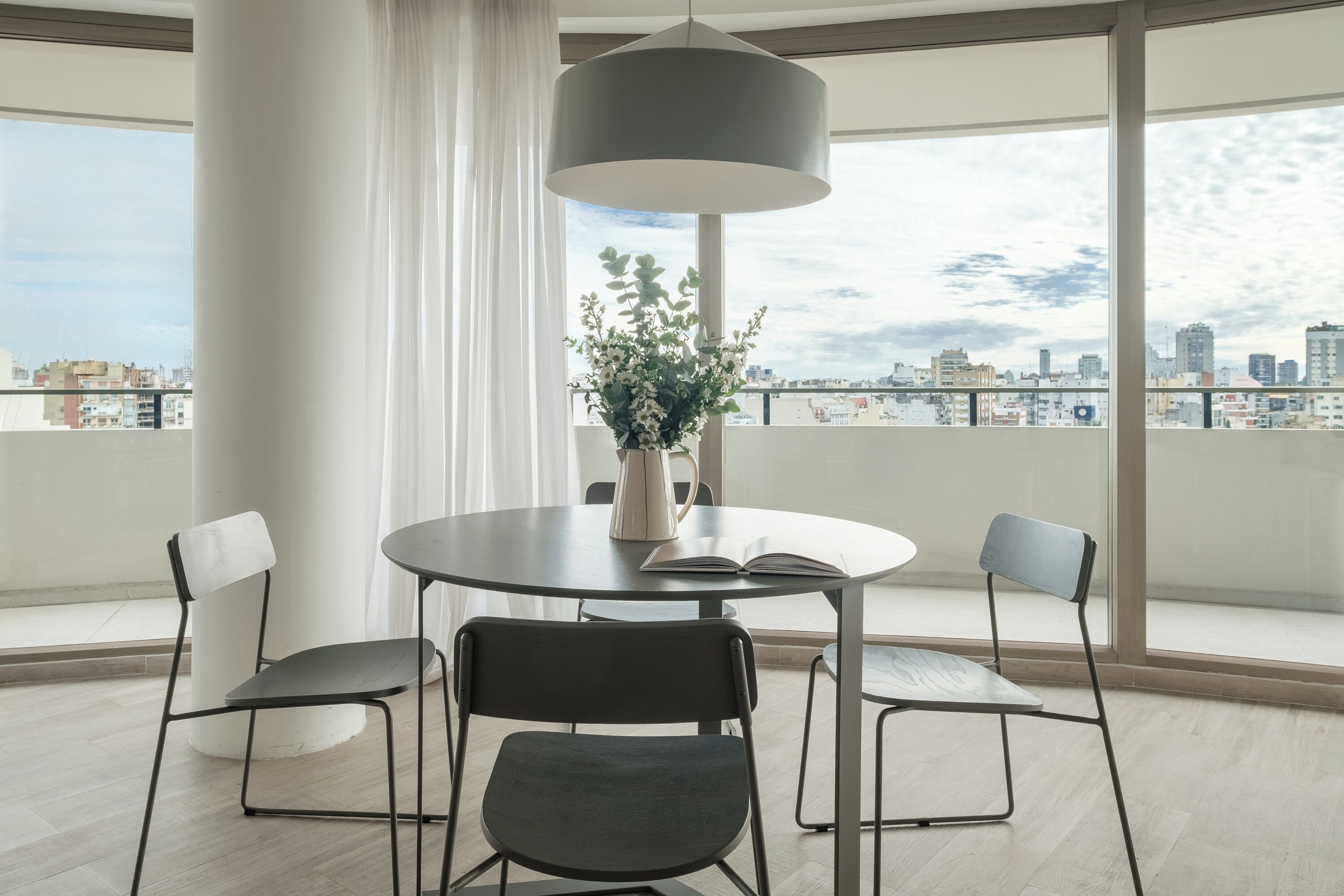 Una habitación moderna con una mesa redonda, cuatro sillas, una planta en maceta y un amplio ventanal con vistas a la ciudad.