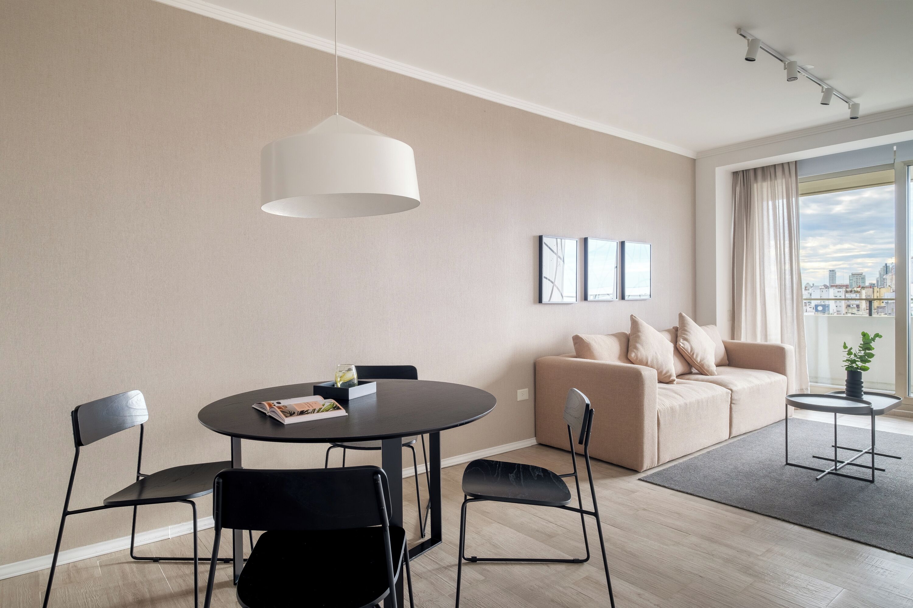 A modern living room with a round dining table, black chairs, a beige sofa, and a city view through large windows.