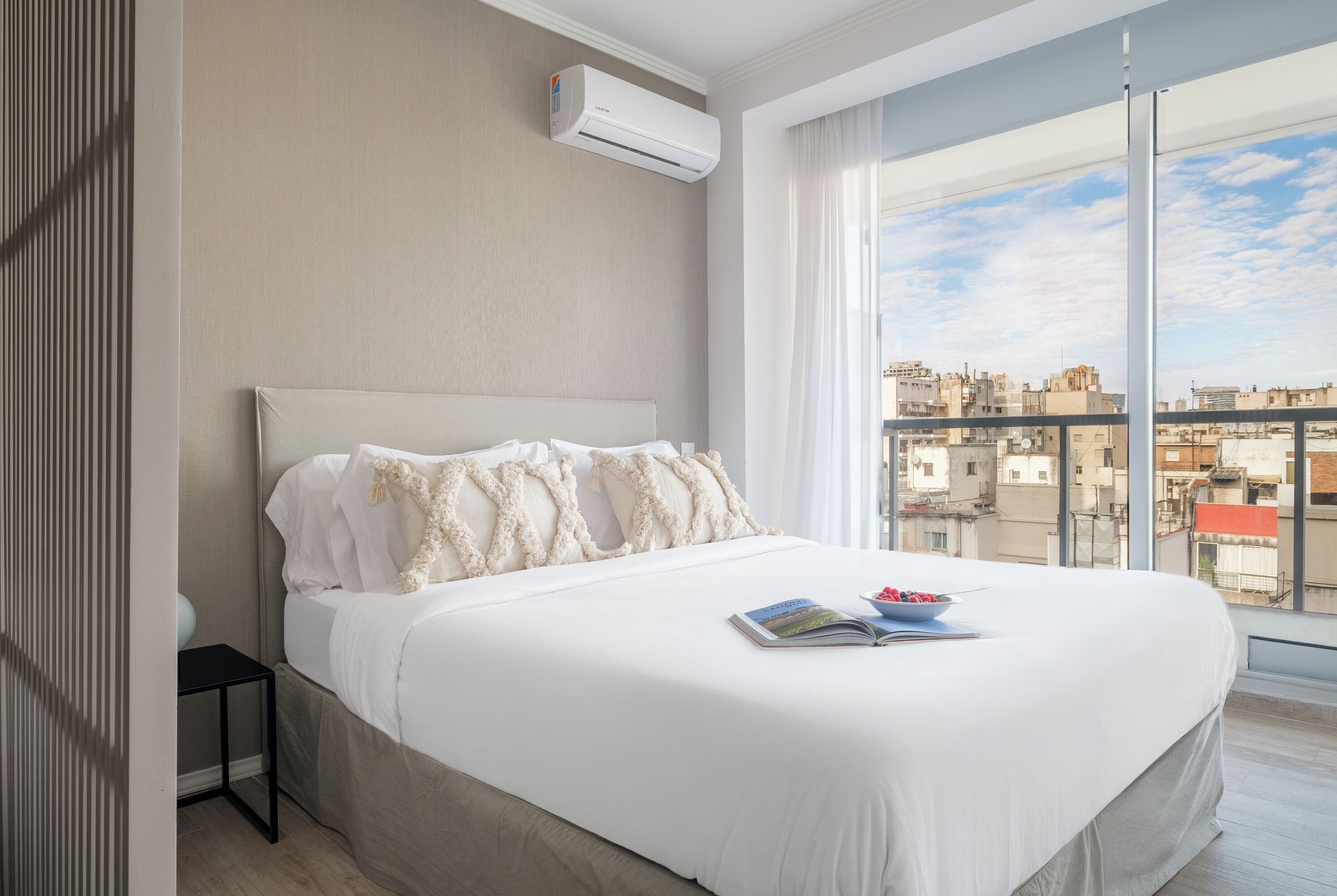 A modern hotel room with a large bed, a small bedside table, a window with a city view, and an air conditioning unit.