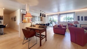 A spacious living area with a dining table, chairs, and a sofa. There are multiple windows providing natural light and a view of the outdoors. - N&J - FLEURS TERRASSE - 3 bedrooms (Niza)