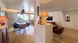 A modern living room with a dining area, a TV mounted on the wall, and a large window providing natural light. - N&J - FLEURS TERRASSE - 3 bedrooms (Niza)