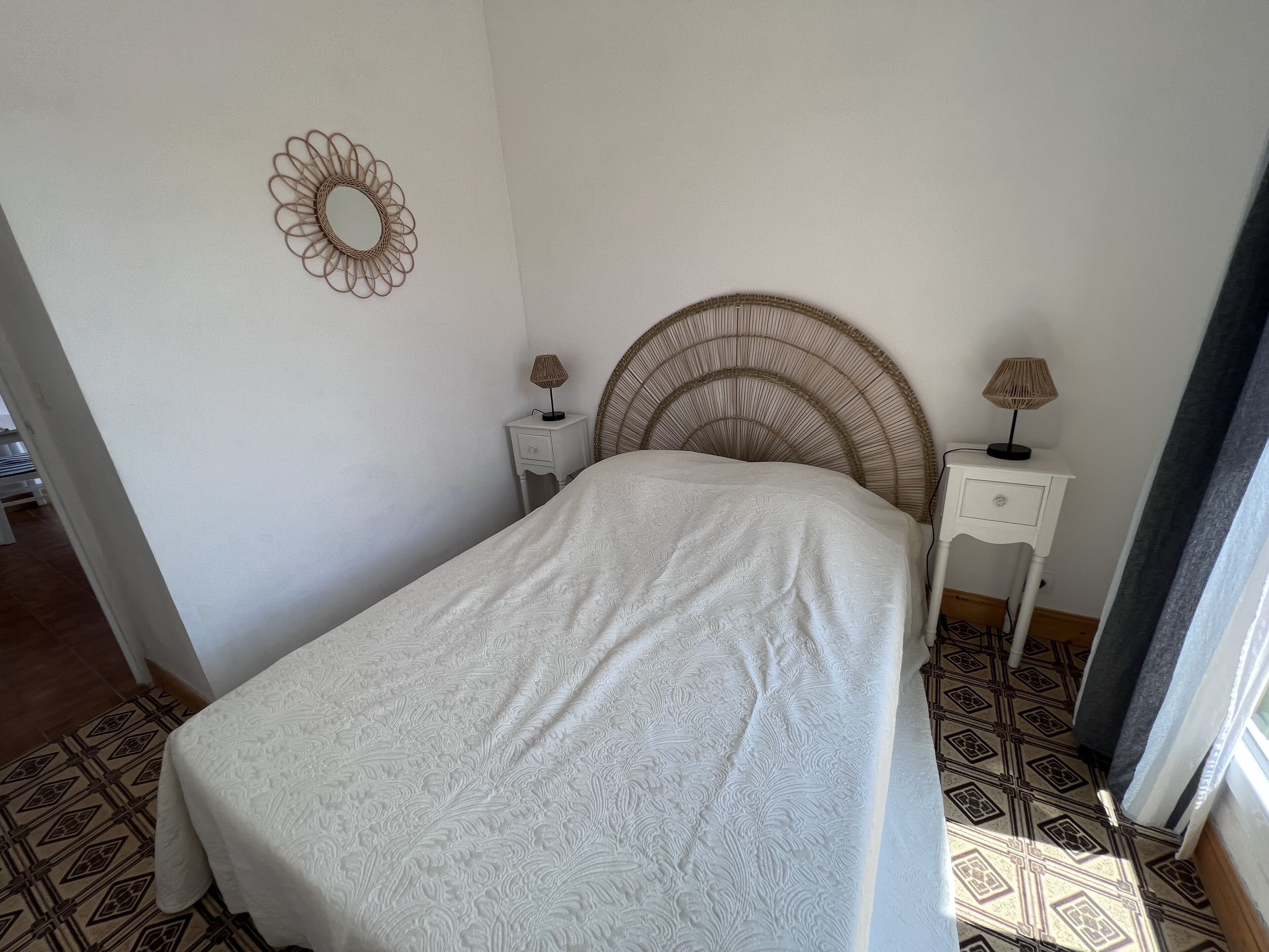 A bedroom with a bed, two bedside tables, a wall-mounted mirror, and patterned flooring.